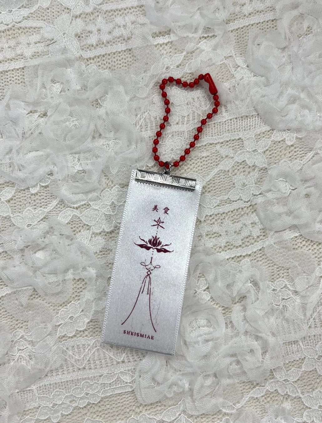白いリボンに「美愛」の文字と花のイラストが描かれたお守り。赤いビーズのチェーンが付いている。背景は白いレース。