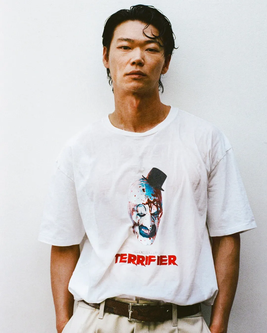 白いTシャツを着た男性が立っている写真。Tシャツには、映画「テリフィアー」のホラーキャラクターのグラフィックがプリントされている。