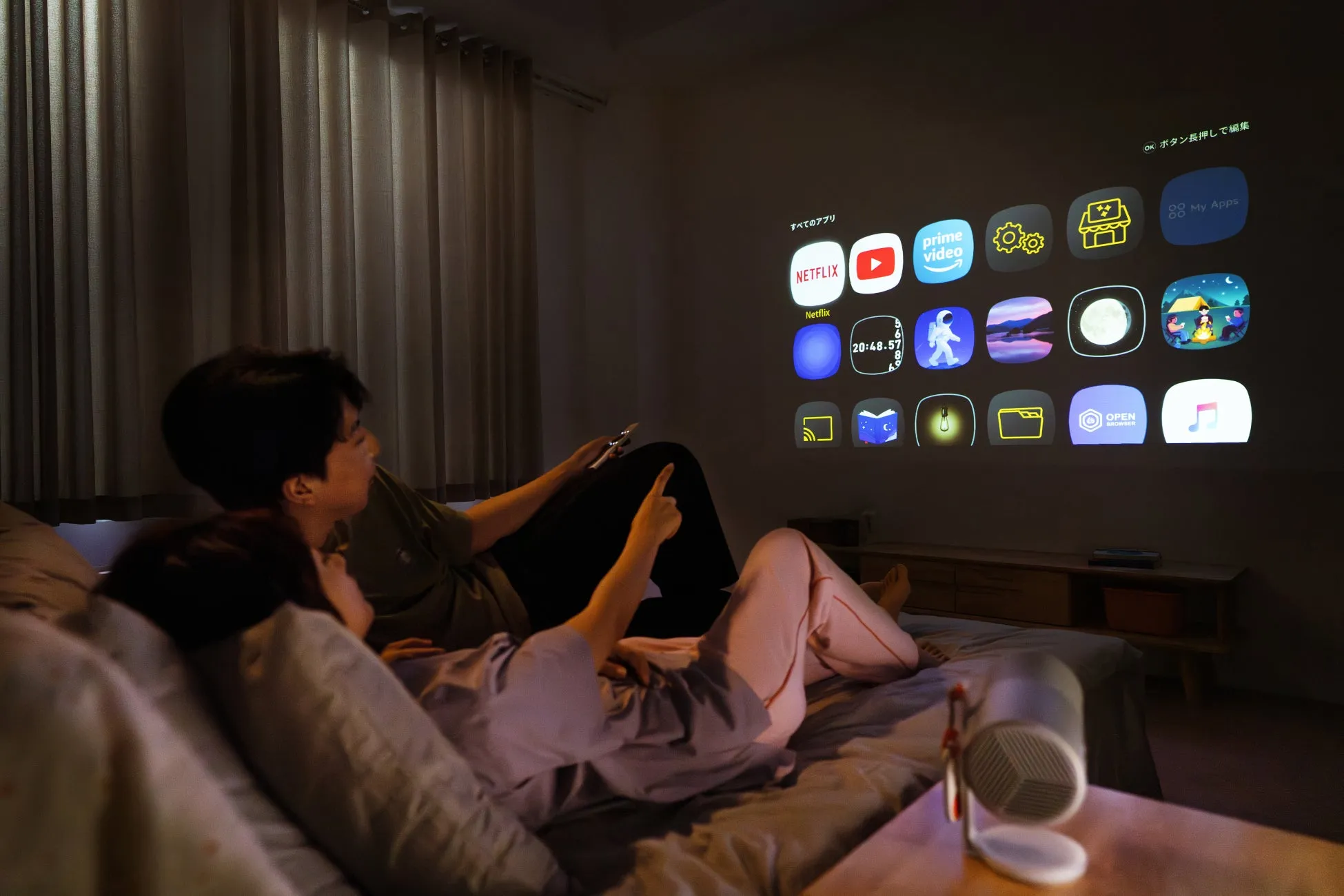 ベッドでくつろぎながらプロジェクターで映像を見ている2人。壁にはアプリのアイコンが表示されており、NetflixやYouTubeなどのサービスが利用できることがわかる。