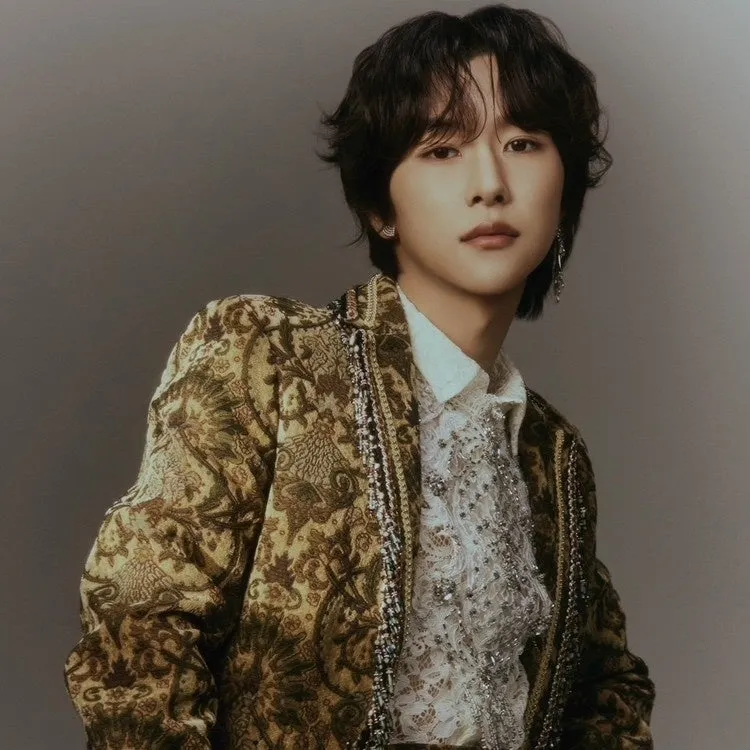 男性が正面を向いて立っており、茶色と金色の模様のジャケットを着ています。彼の肌は白く、黒髪です。彼は白いシャツを着ており、ジャケットはレースのトリムで装飾されています。