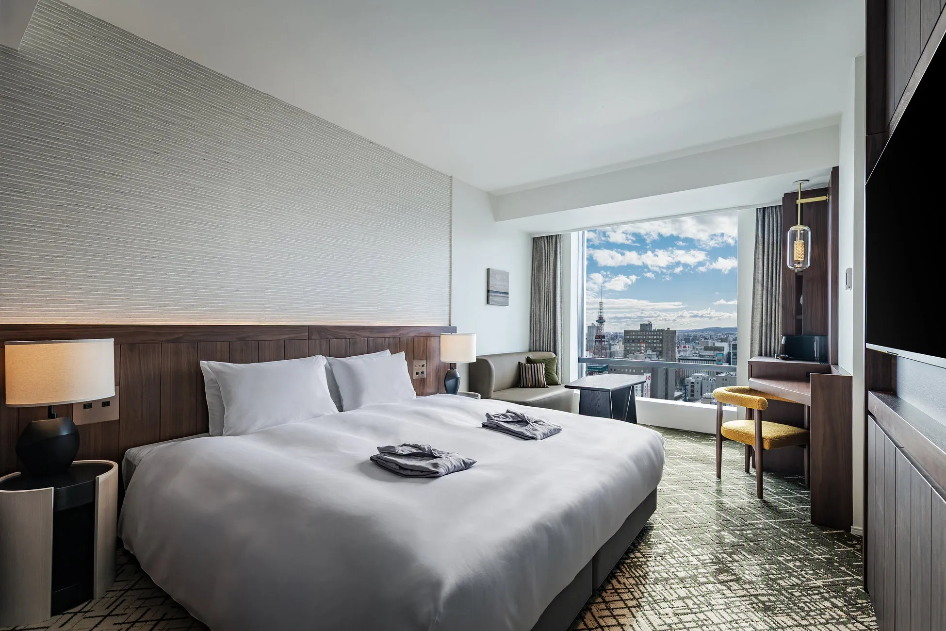 ホテルの客室。ベッド、窓、椅子、テーブル、テレビ、照明が見える。