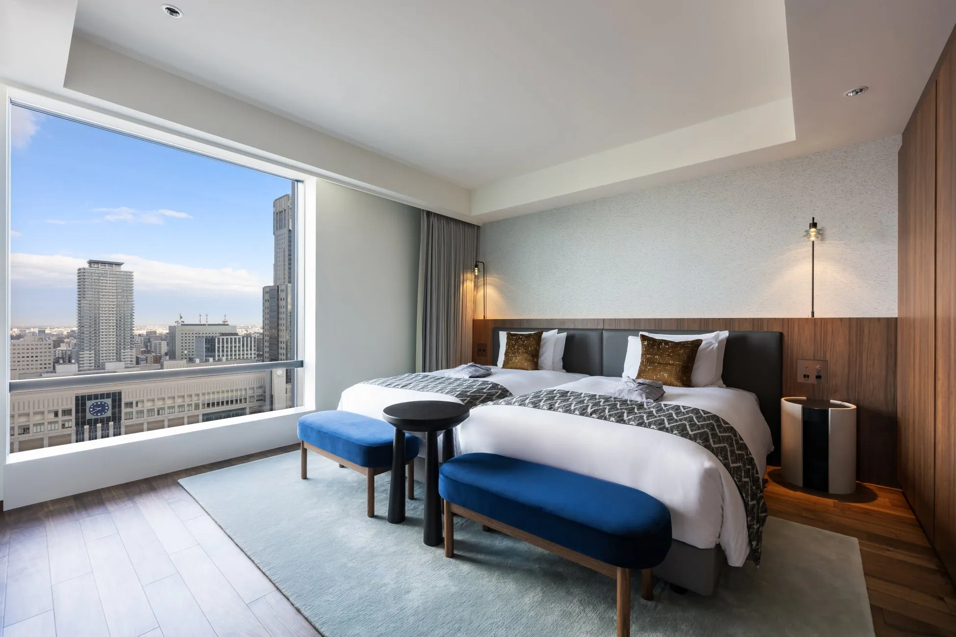 窓から街並みを望むホテルのツインルーム。青いベンチとラグ、黒いサイドテーブルが配置され、洗練された空間を演出している。