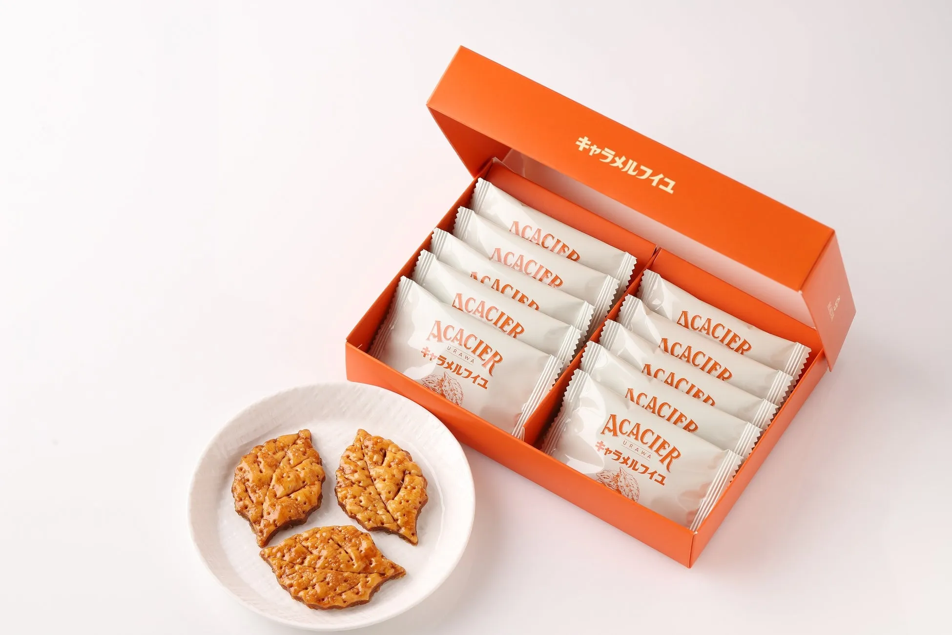 オレンジ色の箱に入ったお菓子。箱には「ACACIER」の文字と「キャラメルフイユ」の文字が書かれている。箱の横には、同じお菓子が白い皿に3つ置かれている。