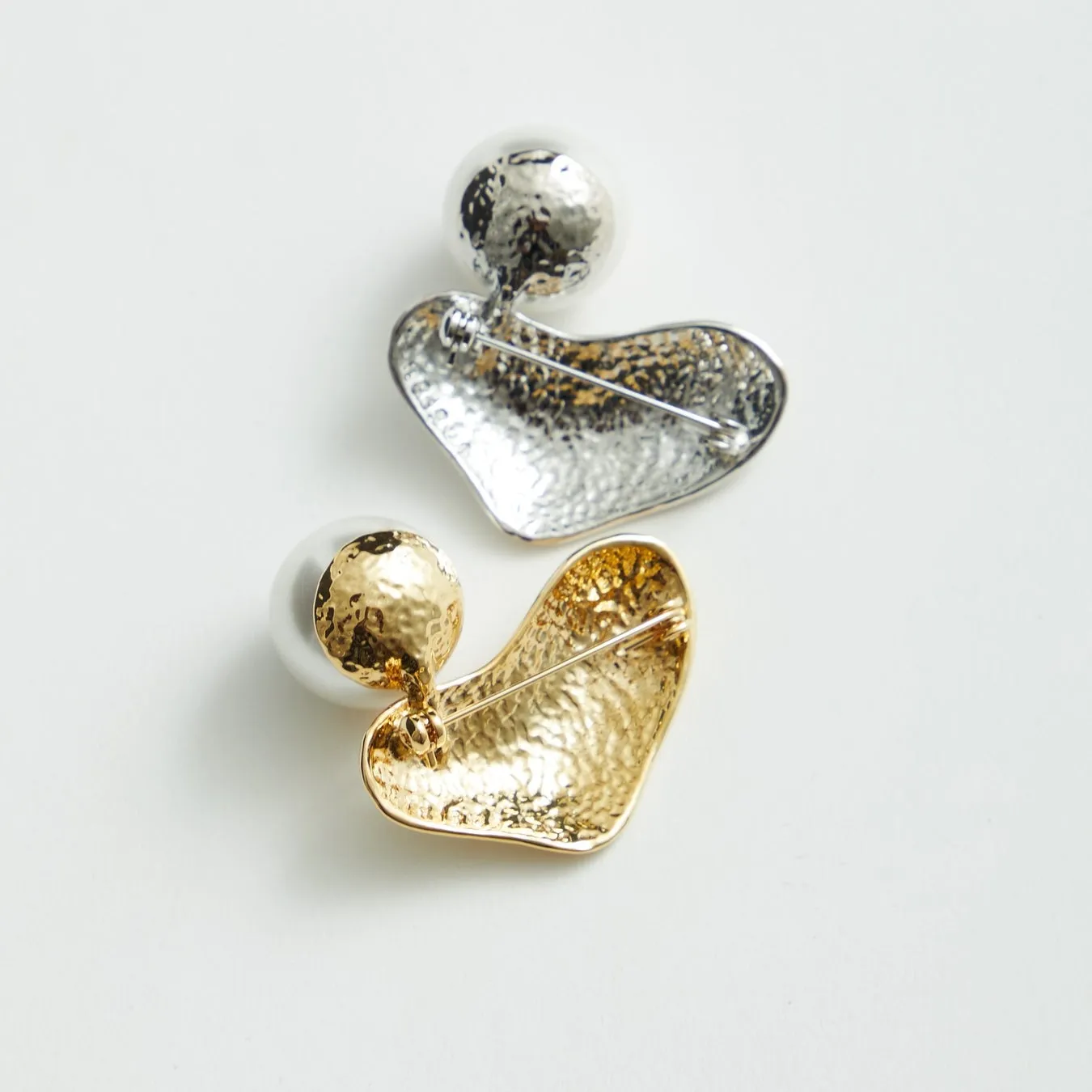 2つのブローチが白い背景に置かれています。上側のブローチは銀色で、ハートの形をしており、丸いアクセントが付いています。下側のブローチは金色で、同じような形をしています。どちらのブローチもピンが付いています。