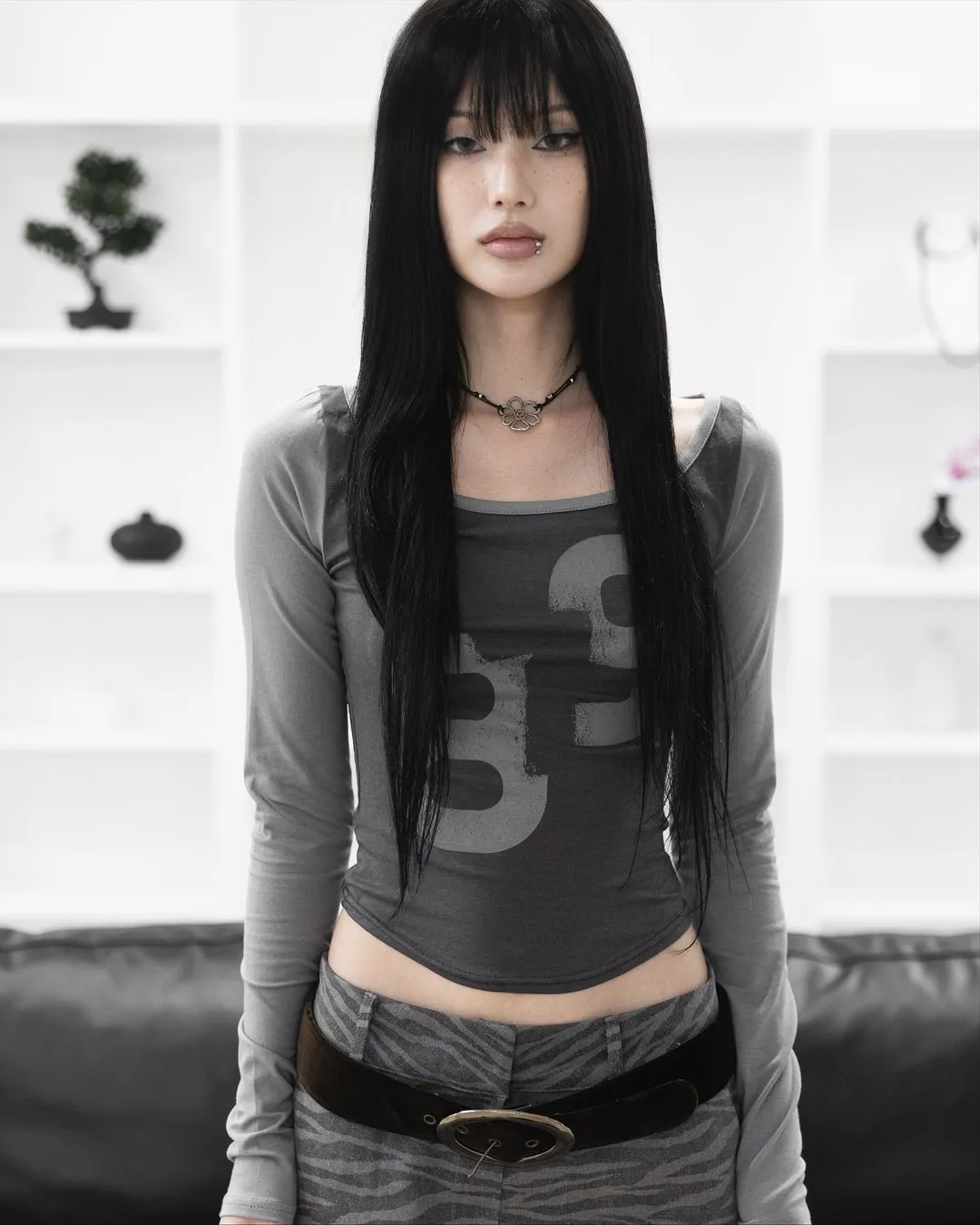 女性が正面を向いて立っており、長くて黒い髪をしていて、前髪があり、唇にピアスをしています。彼女はグレーの長袖シャツと、ウエスト部分にベルトが付いたグレーの模様のパンツを着用しています。背景には白い棚があり、上には小さな盆栽と黒い装飾品があります。