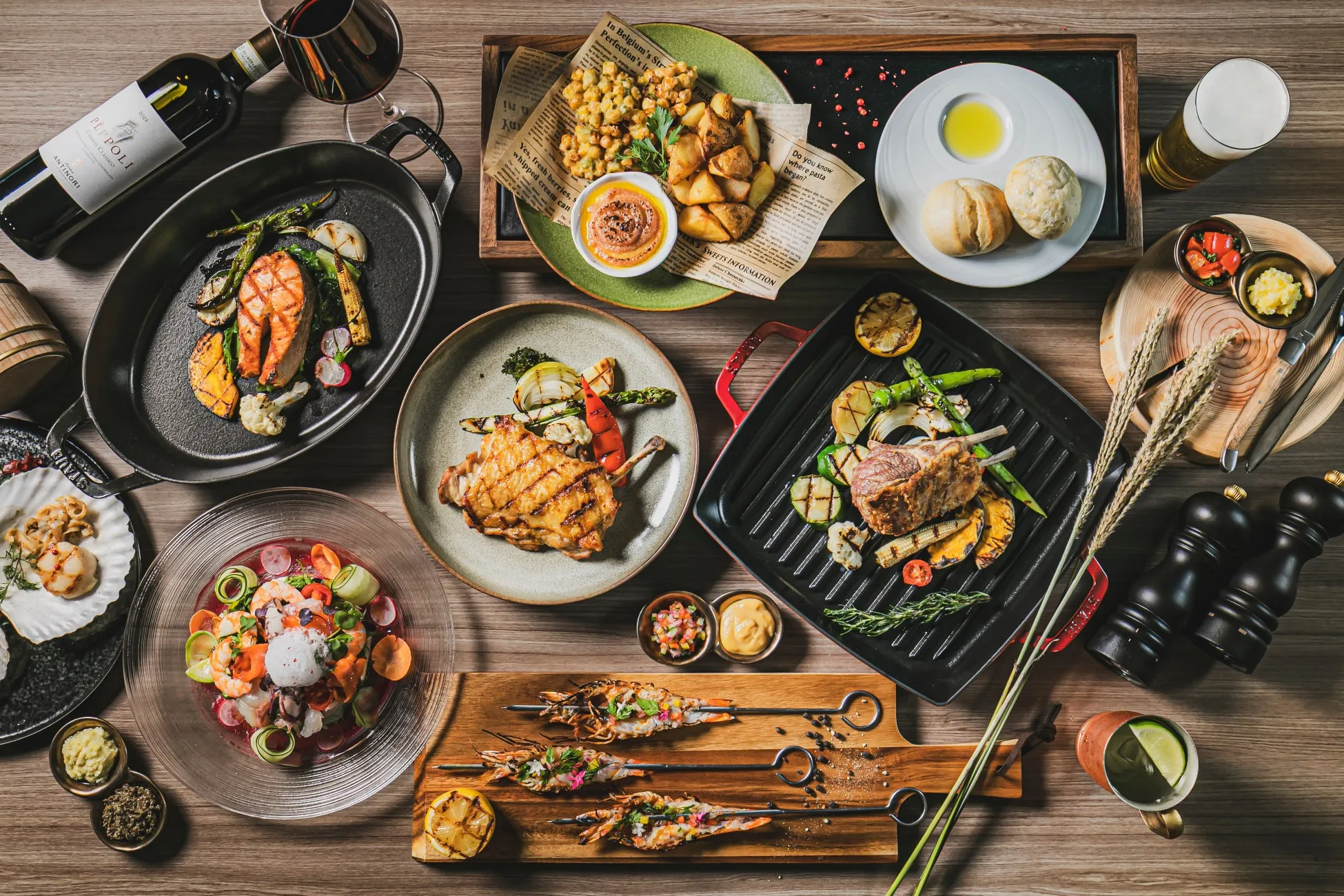 テーブルに並べられた様々な料理の俯瞰写真。グリルされた肉や魚、野菜、エビ、サラダ、パン、ワイン、ビールなど、さまざまな種類の料理が写っています。料理は美しく盛り付けられ、食欲をそそるように見えます。