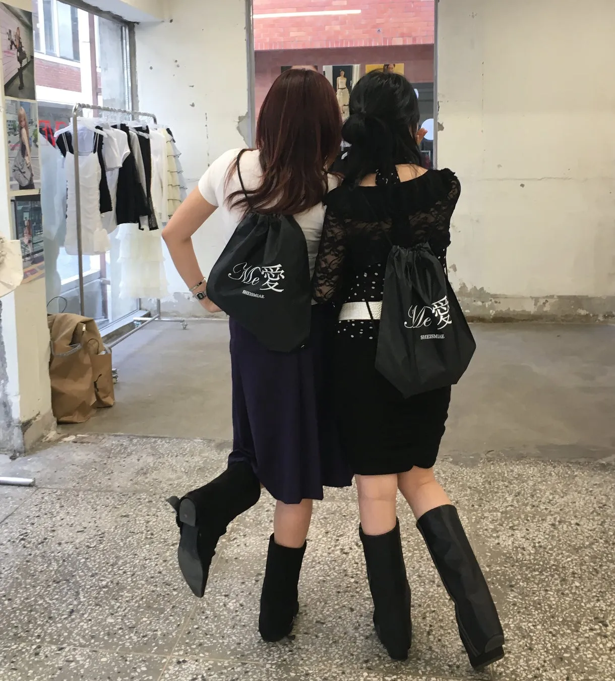 二人の女性が黒い巾着袋を背負い、お互いに寄り添っている様子。背景には、服がかけられたラックとレンガの壁が見える。