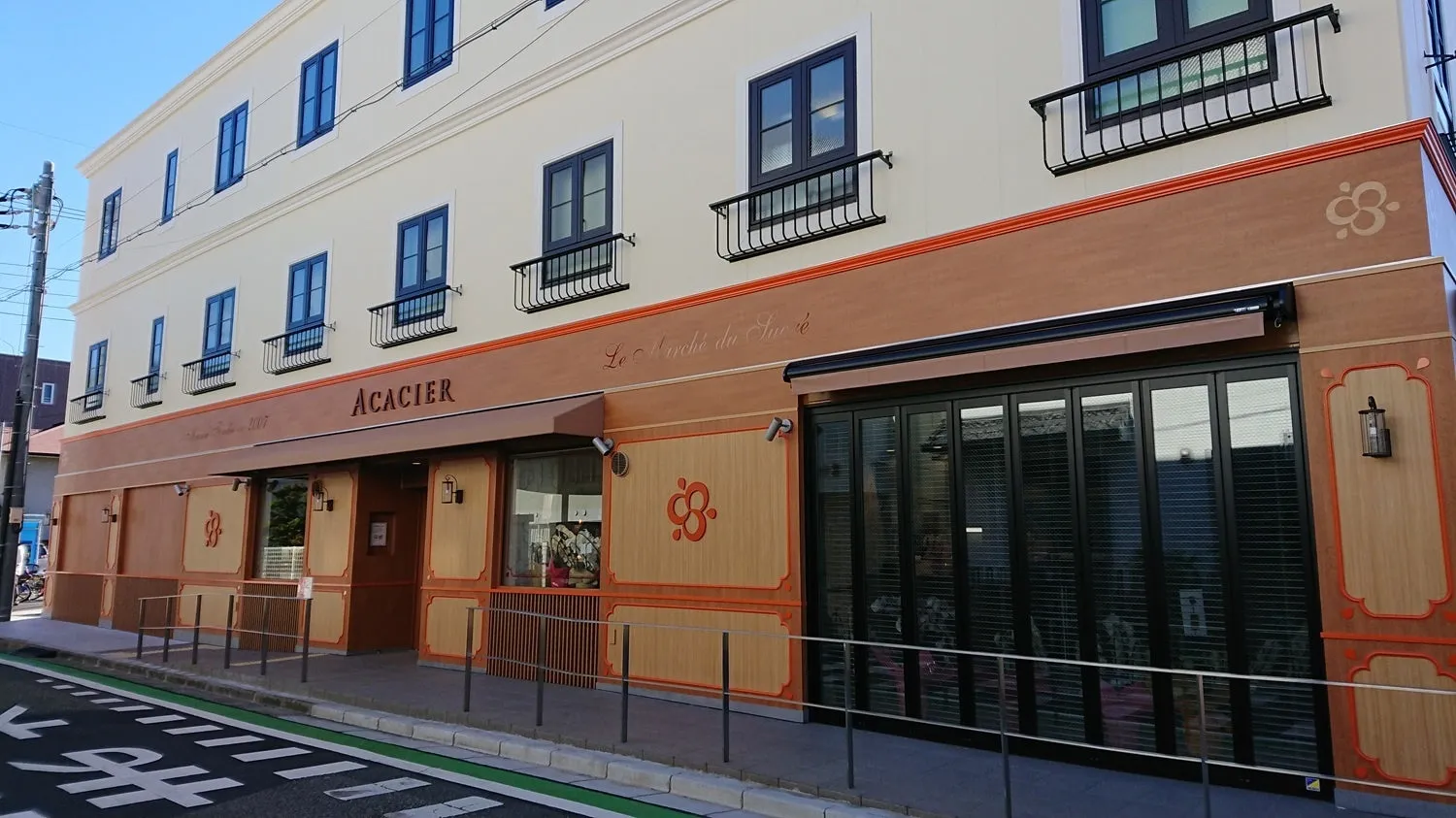 二階建ての建物の外観。正面には「ACACIER」と書かれた看板があり、その下には窓とドアがある。建物はクリーム色と茶色を基調としており、窓には黒い手すりがついている。空は青く晴れ渡っている。