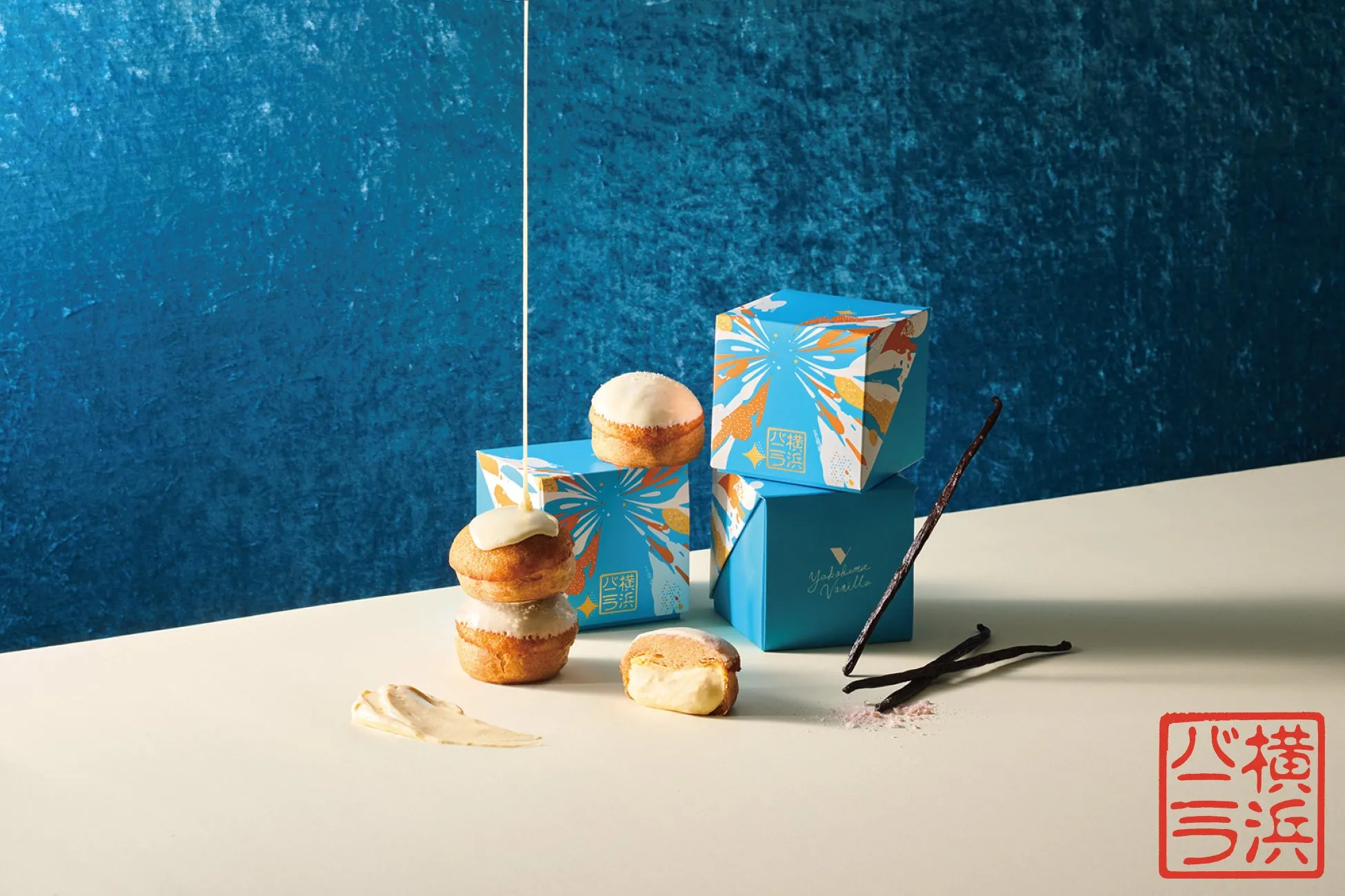 青を基調とした背景に、パッケージされた焼き菓子とバニラビーンズが配置された写真。焼き菓子はクリームでコーティングされており、一部はカットされてクリームが露出している。赤い印鑑のようなものが配置されている。