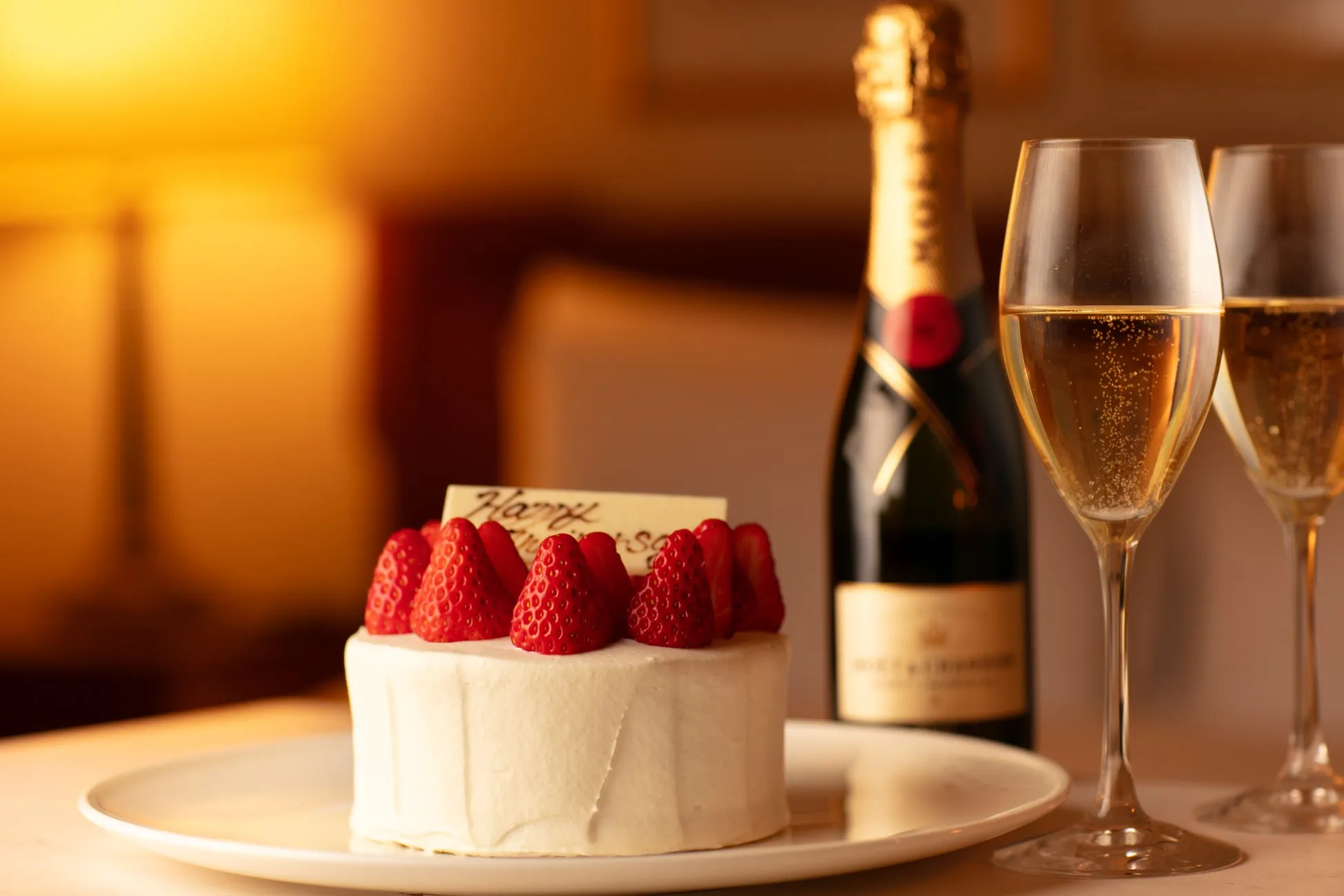 ケーキとシャンパンの入ったグラスがテーブルに置かれた写真です。ケーキにはイチゴがトッピングされており、Happyの文字が書かれたチョコレートプレートが添えられています。シャンパンのボトルも写っています。