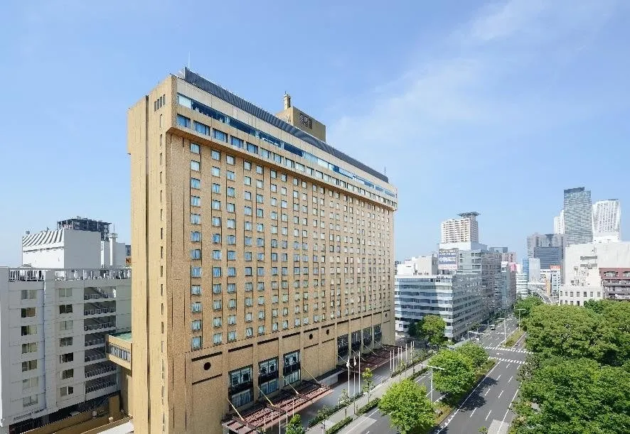 高層ホテルの建物の外観を捉えた写真です。建物は茶色で、多くの窓があり、空は青く晴れています。ホテルの前には道路と並木があり、街並みが広がっています。