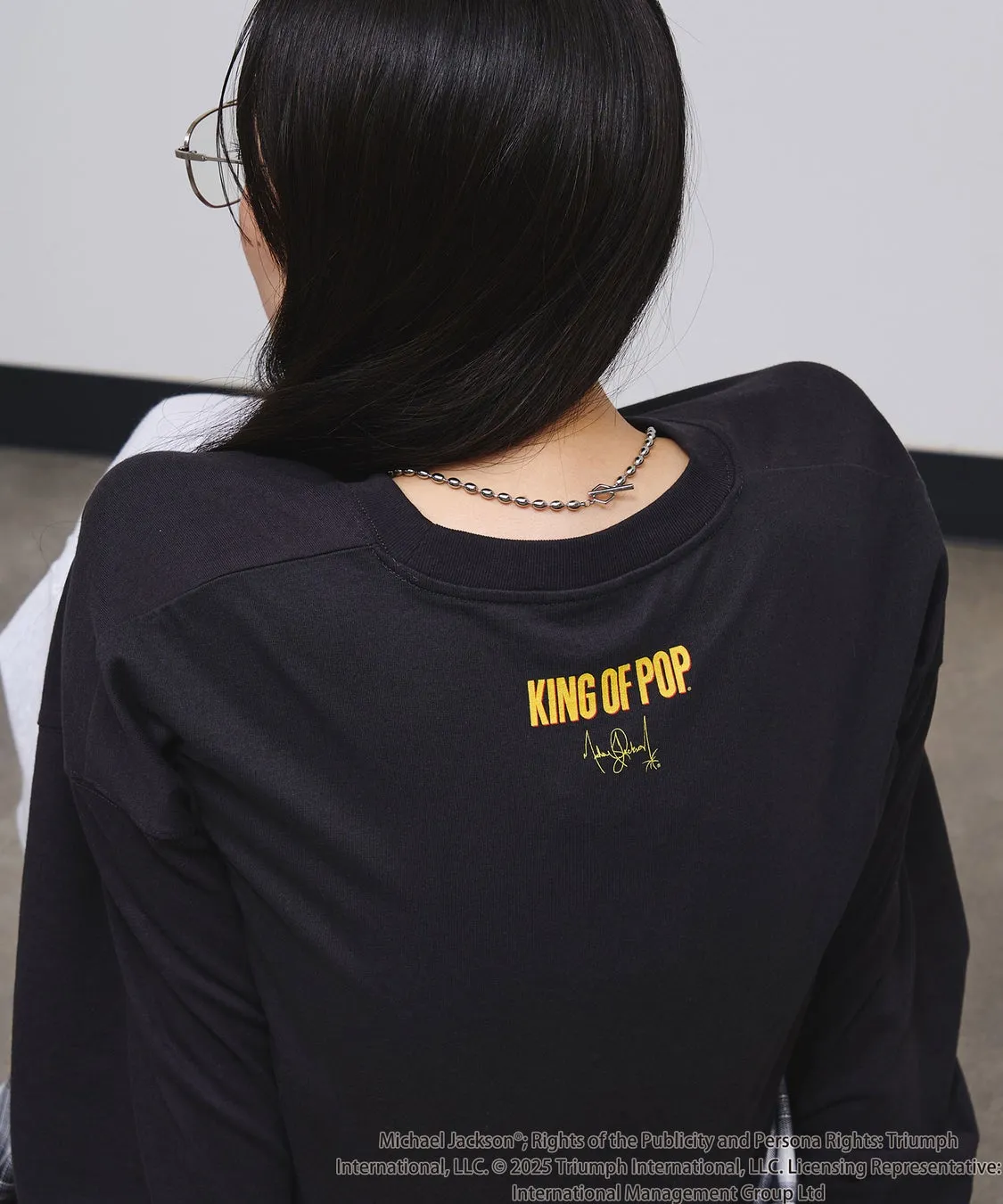 後ろ姿の女性が写っており、黒い長袖の服を着ています。服には「KING OF POP」という文字とサインがプリントされています。