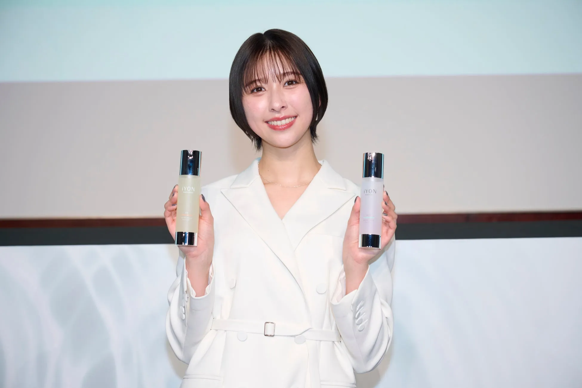 白い服を着た女性が、化粧品のボトルを2つ持って微笑んでいる写真