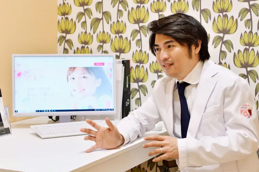 白衣を着た男性が、パソコンの画面を見ながら話をしている様子。背景には花柄の壁紙がある。