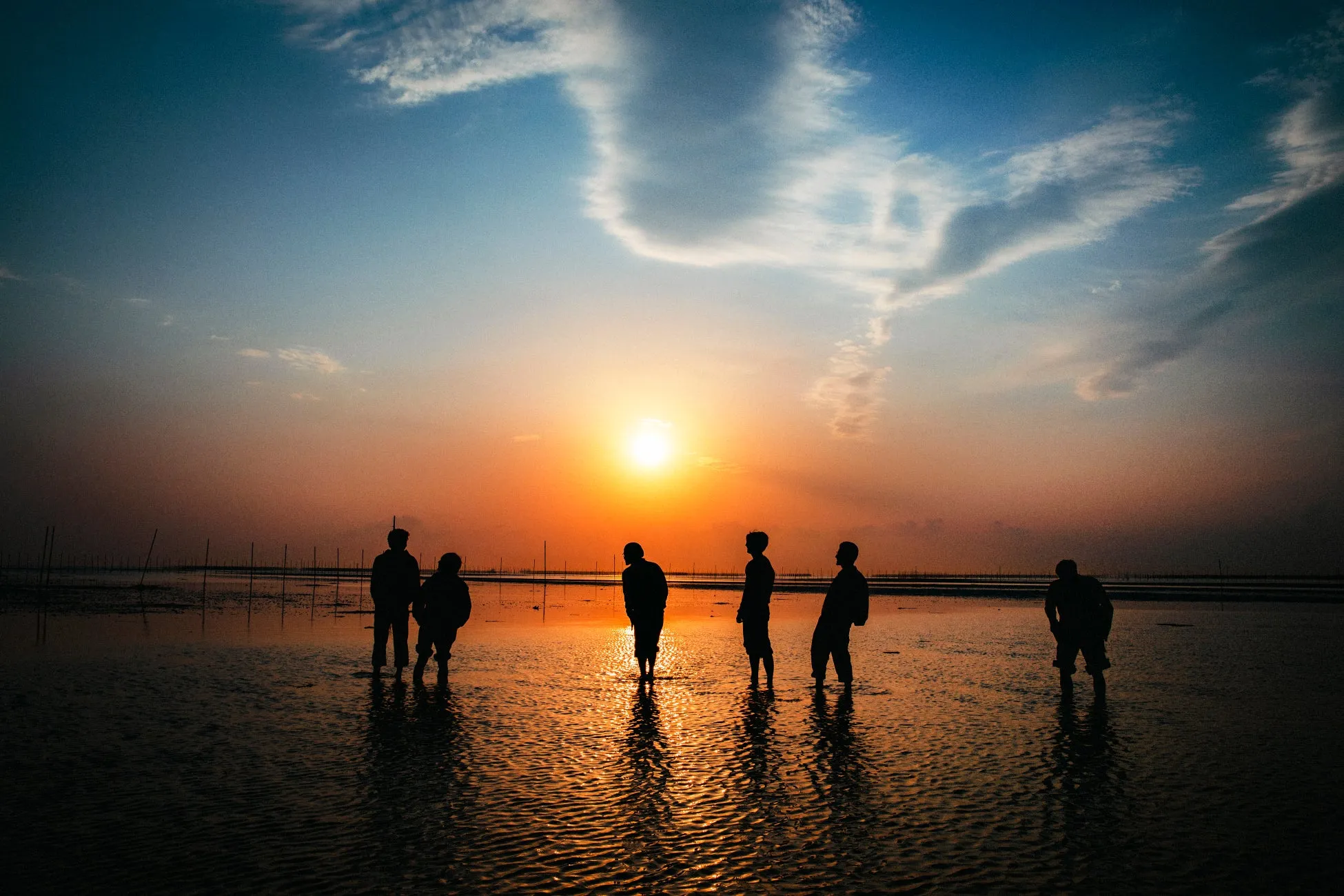 夕焼け空の下、水面に人が立っているシルエットを捉えた写真。オレンジ色の夕日が水面に反射し、美しい光景を作り出している。人々は水遊びをしているようにも見える。