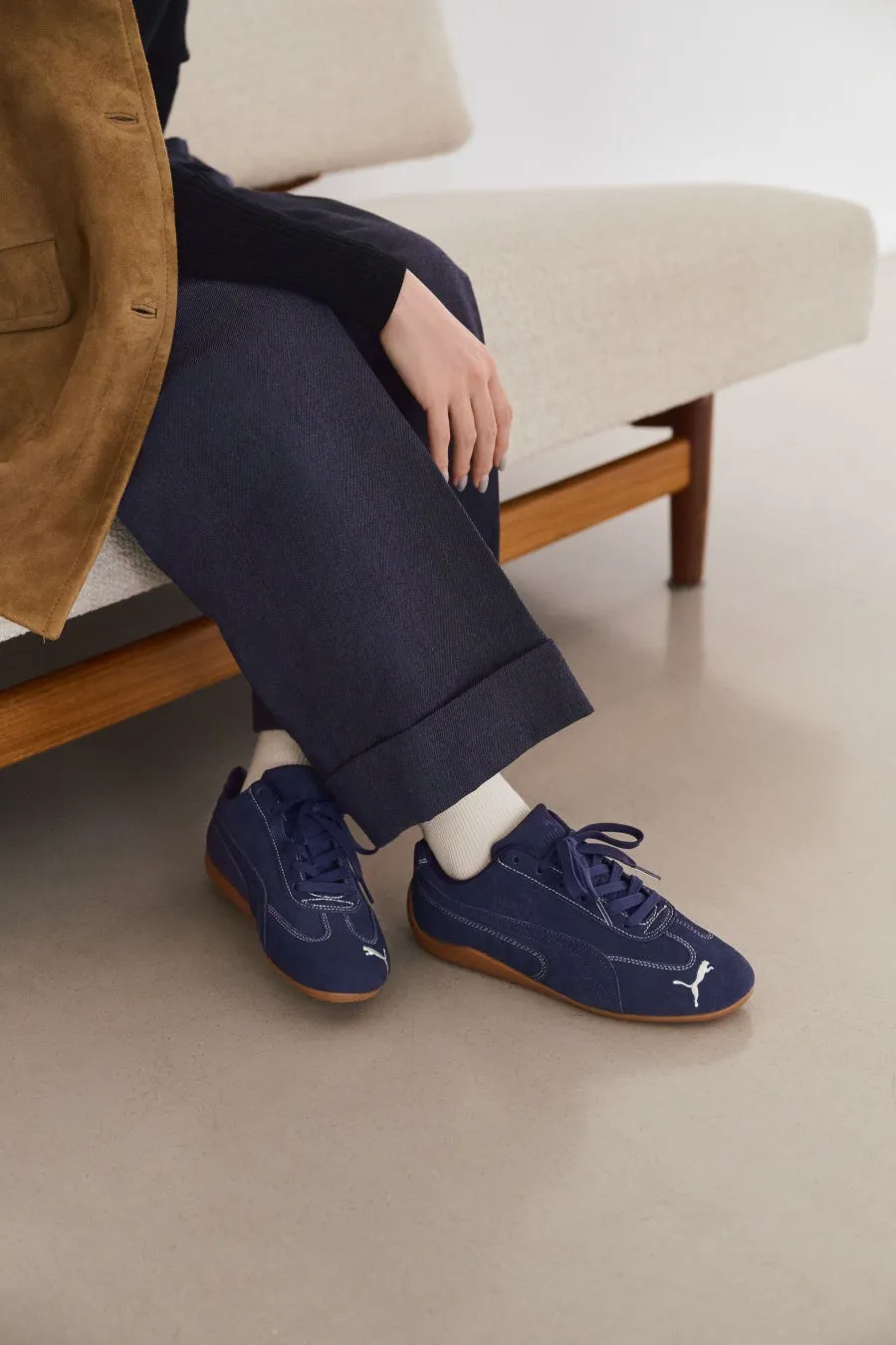 デニム素材のプーマのスニーカーを履いた女性。デニムのパンツを履いており、白い靴下と茶色のソールが見える。背景には椅子がある。