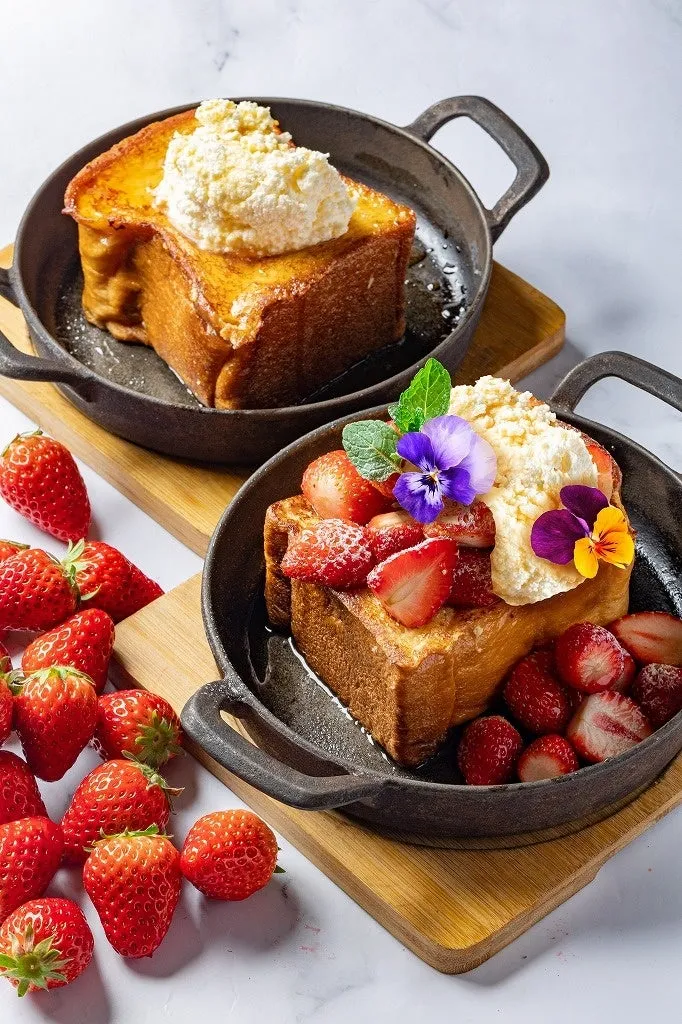 2つのパンケーキが、それぞれ異なるトッピングで提供されている様子。1つはクリームがトッピングされ、もう1つはイチゴと花がトッピングされています。イチゴが散りばめられており、背景は白です。