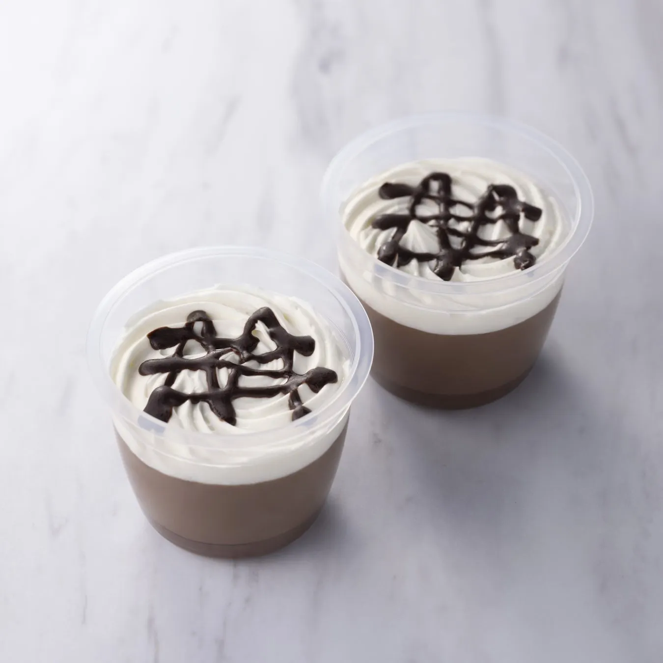 2つのチョコレートムースのデザートが、白いクリームとチョコレートソースでトッピングされ、プラスチックの容器に入っています。大理石のテーブルの上に置かれています。