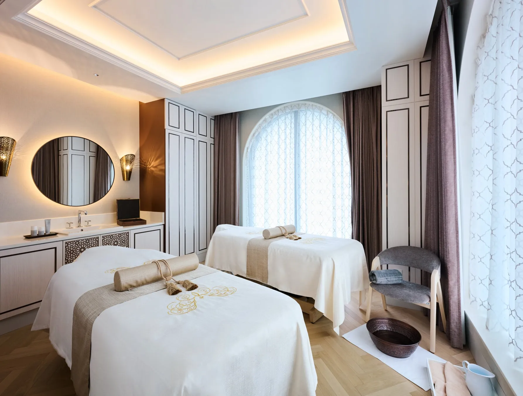 スパルームのインテリア。2つのマッサージベッド、丸い鏡、椅子、フットバス、窓、カーテンが写っています。