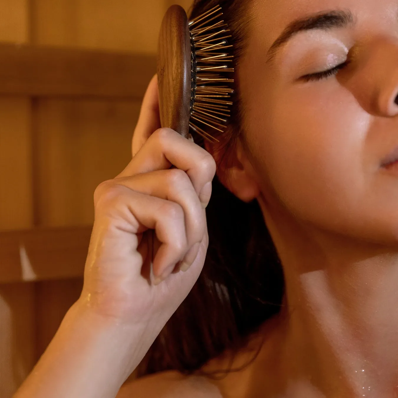 女性が髪をとかしている様子。木製のブラシと、閉じた目、リラックスした表情が写っている。