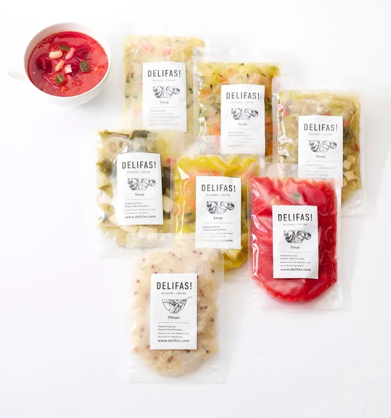 様々な種類のスープと粥が透明な袋に入っており、白い背景に配置されている写真です。左上にはスープが入った白い器があります。