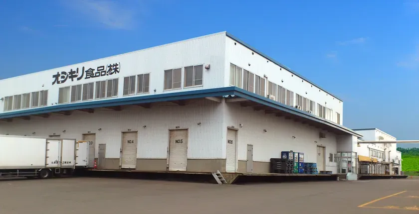 オシキリ食品株式会社の倉庫の外観写真。白い建物で、複数の搬入口があります。空は青く晴れています。