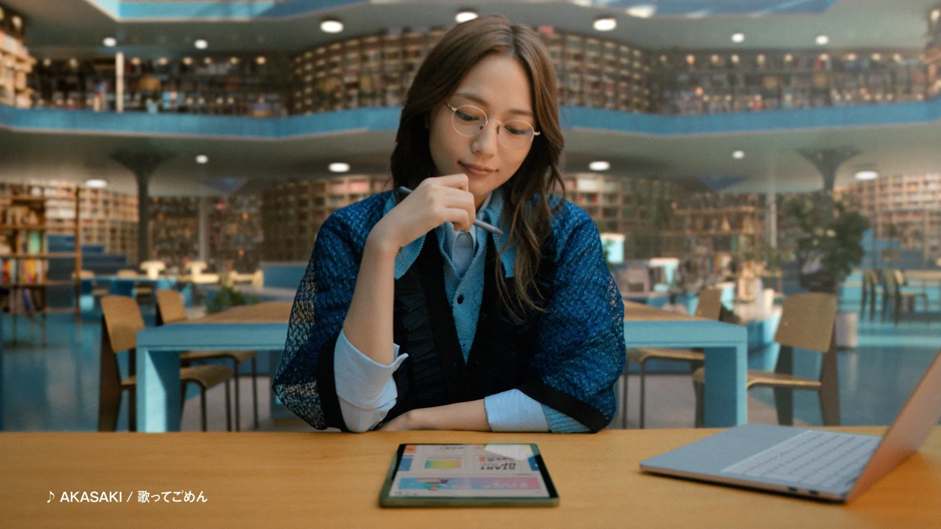 女性が図書館のような場所でタブレットとノートパソコンを使って作業をしている様子を描いた画像です。女性は眼鏡をかけ、ペンを持ちながらタブレット画面を見つめています。背景には本棚が並び、落ち着いた雰囲気です。