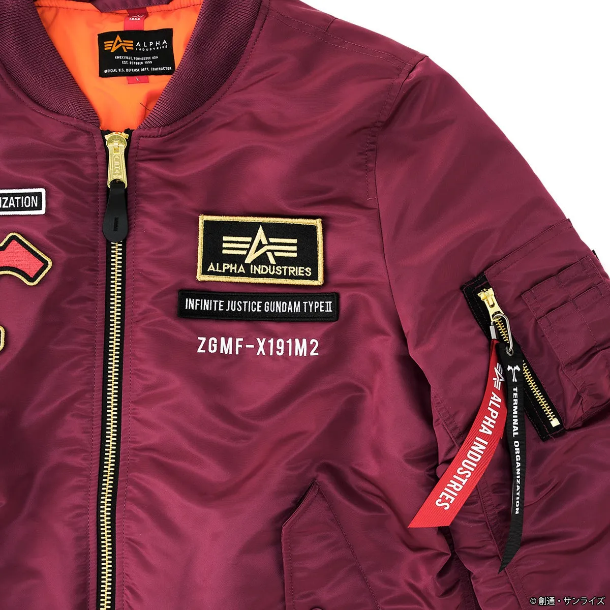 赤色のMA-1ジャケット。左胸にALPHA INDUSTRIESのロゴワッペンとINFINITE JUSTICE GUNDAM TYPEIIの文字が刺繍されたワッペンが縫い付けられている。右袖にはALPHA INDUSTRIESのロゴが入ったタグが付いている。
