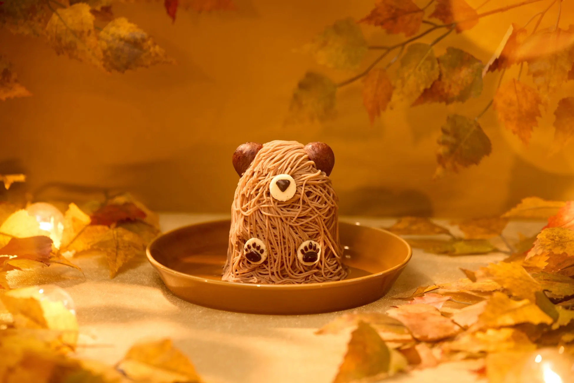 秋の装飾が施された、クマの形をしたモンブランケーキが皿の上に置かれています。背景には紅葉した葉が散りばめられています。