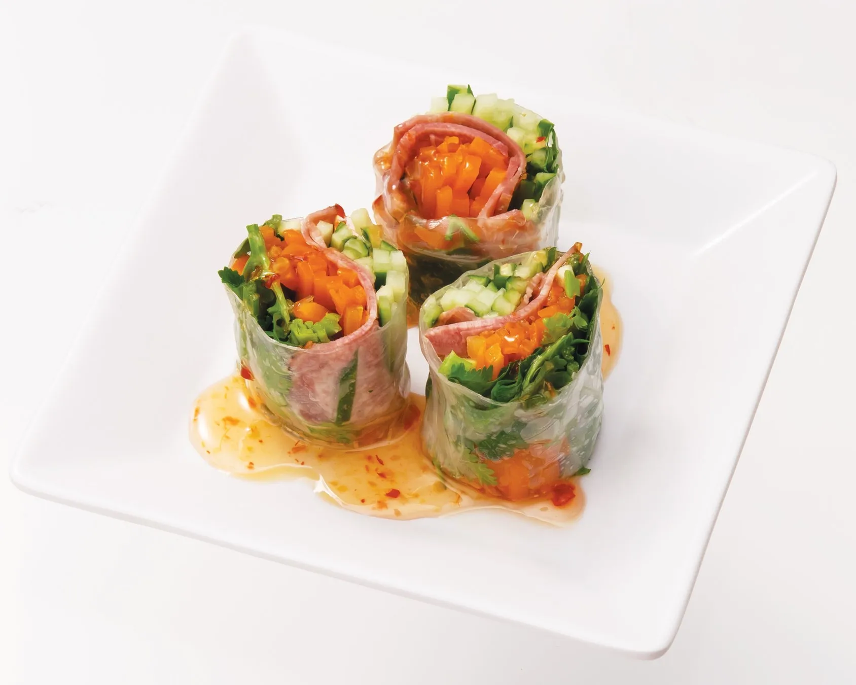 白い皿の上に、3つの春巻きが乗っている写真。春巻きは透明なライスペーパーで包まれ、中にはニンジン、キュウリ、葉物野菜、ハムのようなものが入っている。春巻きの周りにはソースが添えられている。