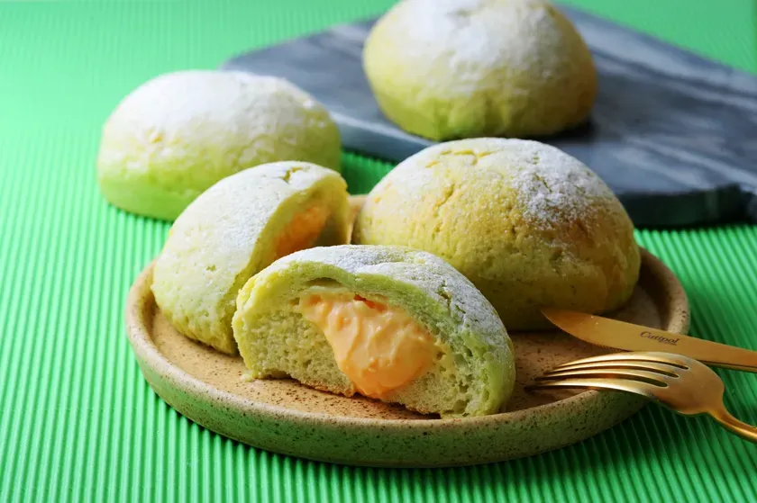 抹茶色のメロンパンの中にオレンジ色のクリームが入っている。皿に盛り付けられ、金色のナイフとフォークが添えられている。緑色の背景に置かれている。