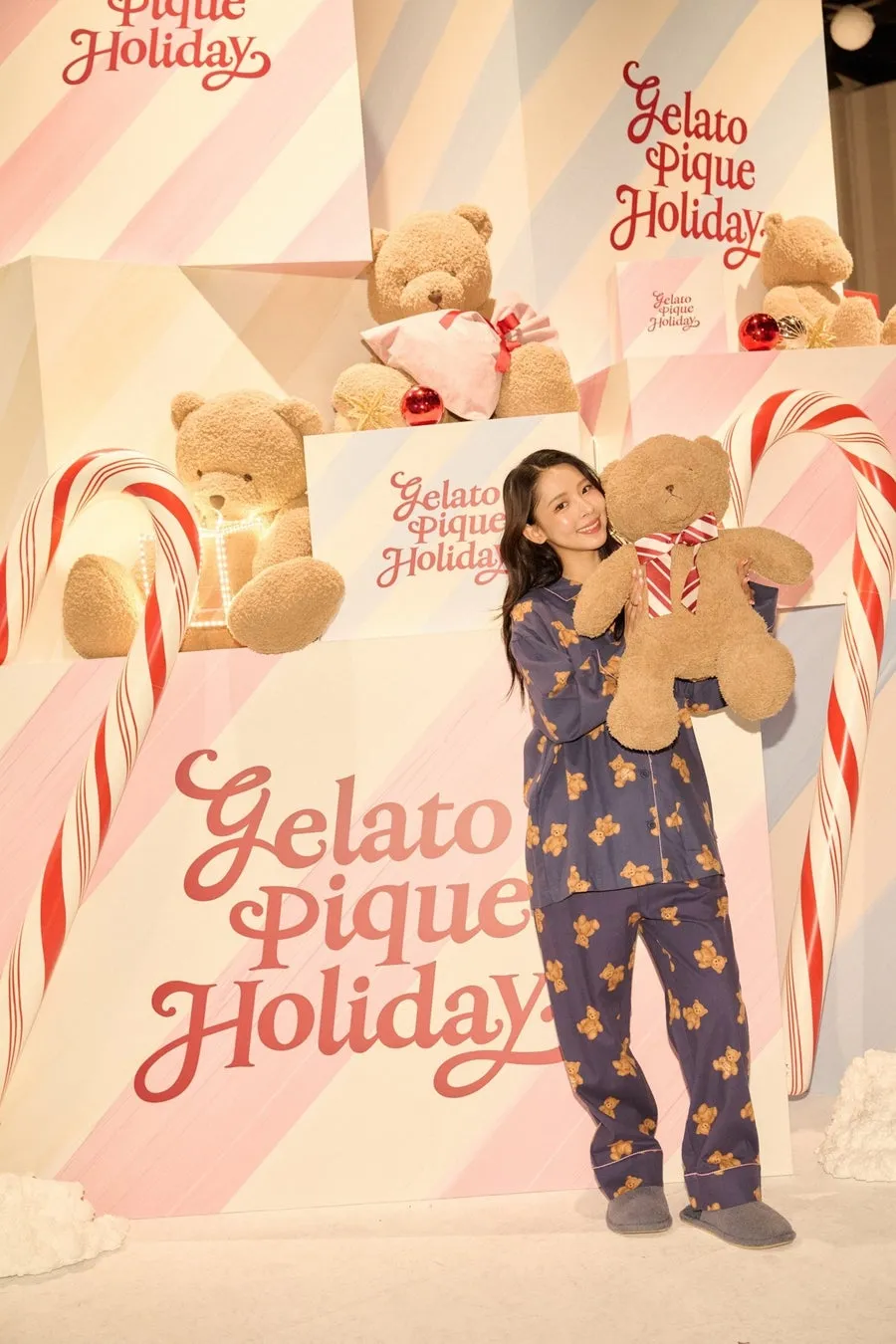 女性がテディベアのぬいぐるみを持って立っており、背景には「Gelato Pique Holiday」のロゴとテディベアの装飾が施されています。