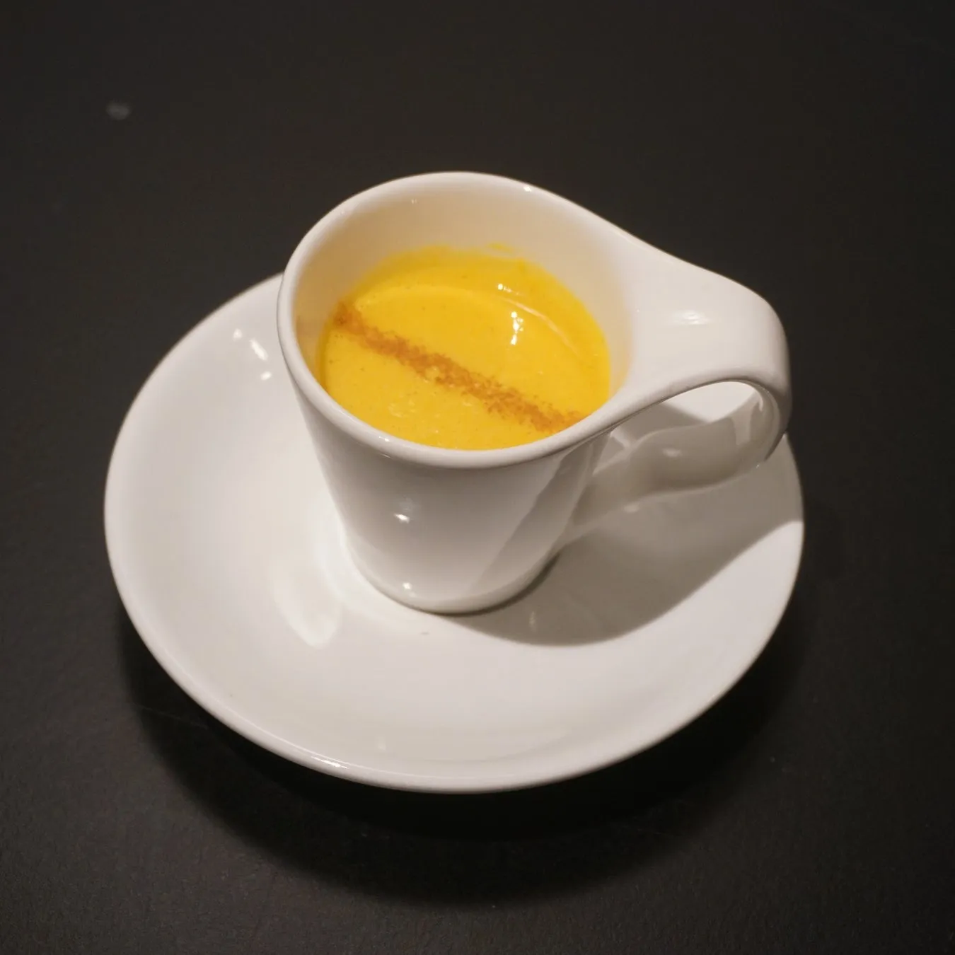 白いカップに入った黄色いスープ。スープの上にはシナモンのようなものがかかっている。カップはソーサーの上に置かれている。
