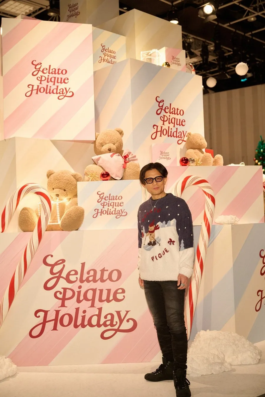 男性が、クリスマスをテーマにした装飾の前で立っている写真。背景には、クマのぬいぐるみ、キャンディケイン、ブランドロゴが配置されています。
