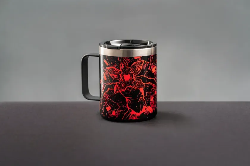黒いマグカップ。マグカップには赤い模様でデモゴルゴンが描かれています。マグカップの取っ手は黒色で、背景はグレーです。