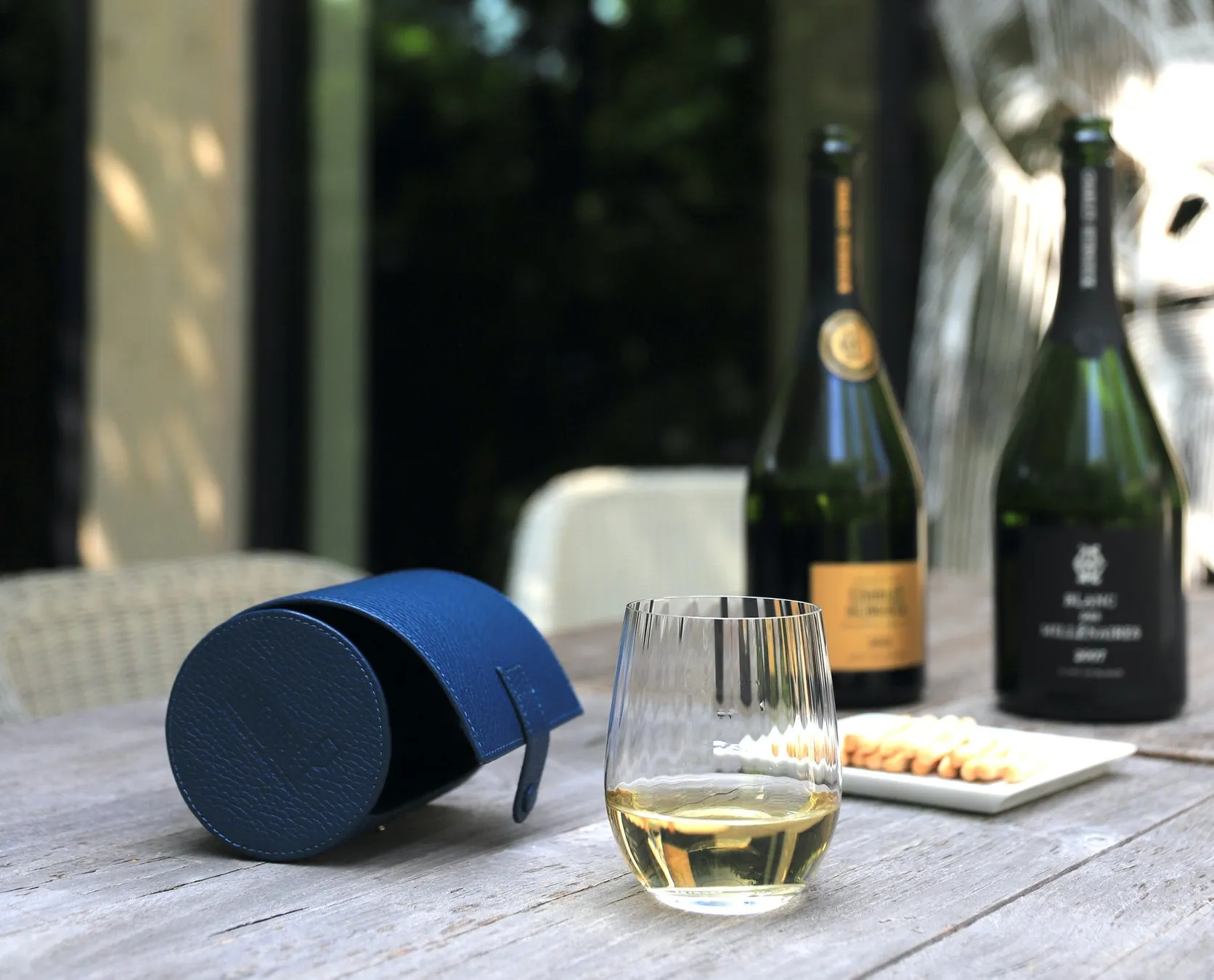 テーブルの上に、ワインボトル、ワイングラス、青い革のワインバッグ、クラッカーが置かれている写真。背景には木々が見える。