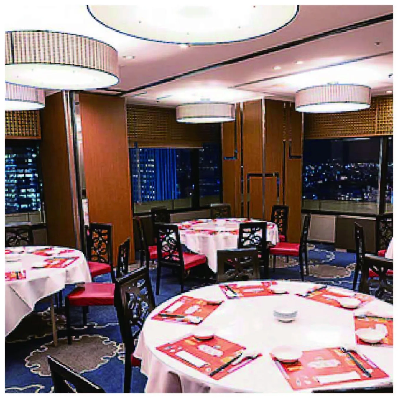 円卓と椅子が並んだレストランの室内写真。テーブルクロスは白で、赤いランチョンマットが置かれている。窓からは夜景が見える。