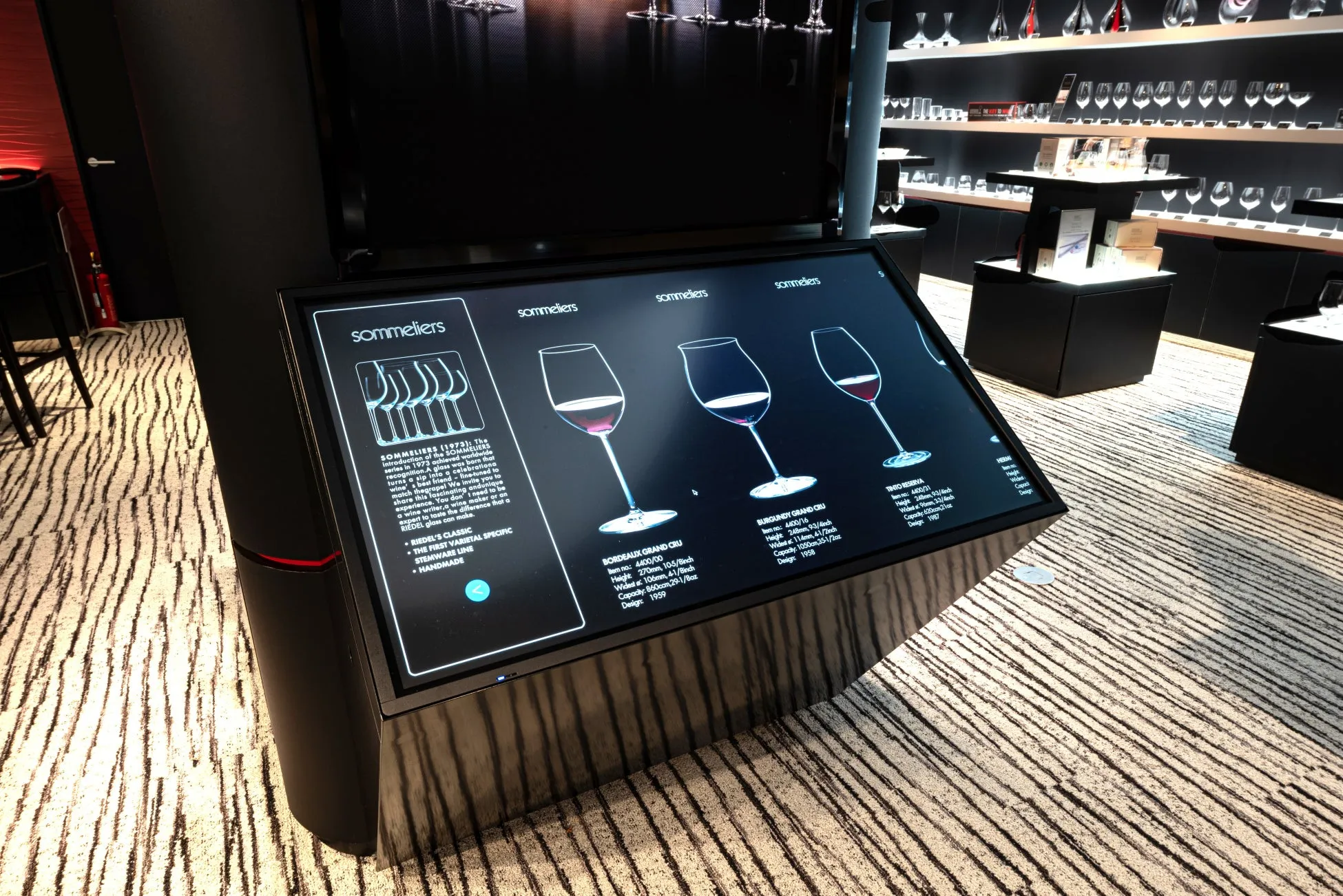 ワイングラスの展示。タッチパネルには、様々な種類のワイングラスの特徴と、それらに適したワインの種類が説明されている。背景にはワイングラスが陳列された棚が見える。