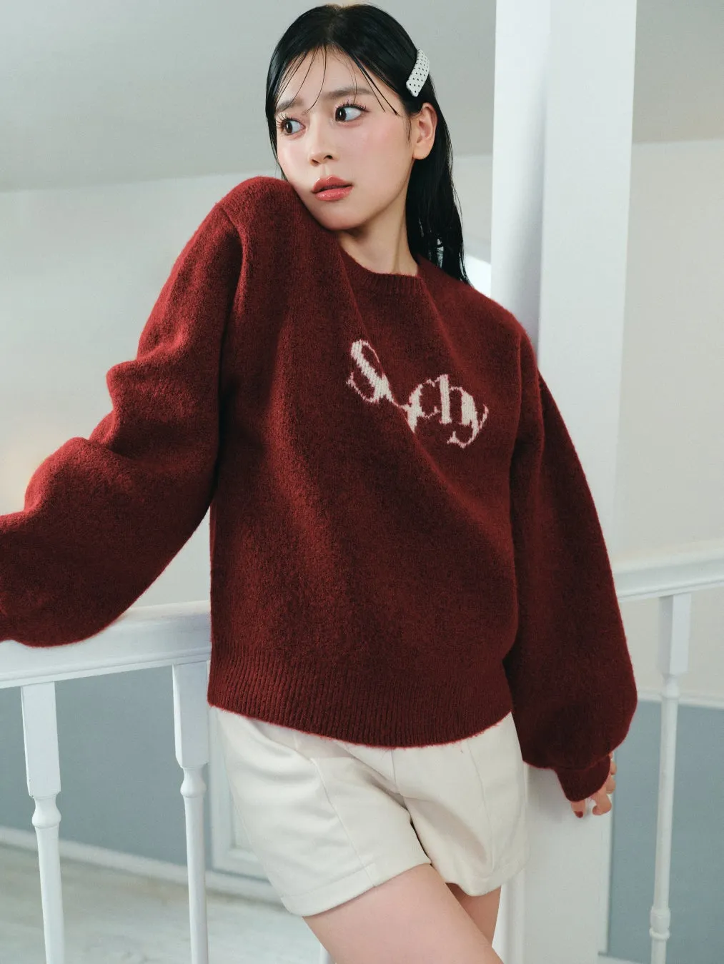 女性が赤いセーターと白いショートパンツを着て、手すりに寄りかかっている写真です。セーターには「Say」の文字がプリントされています。