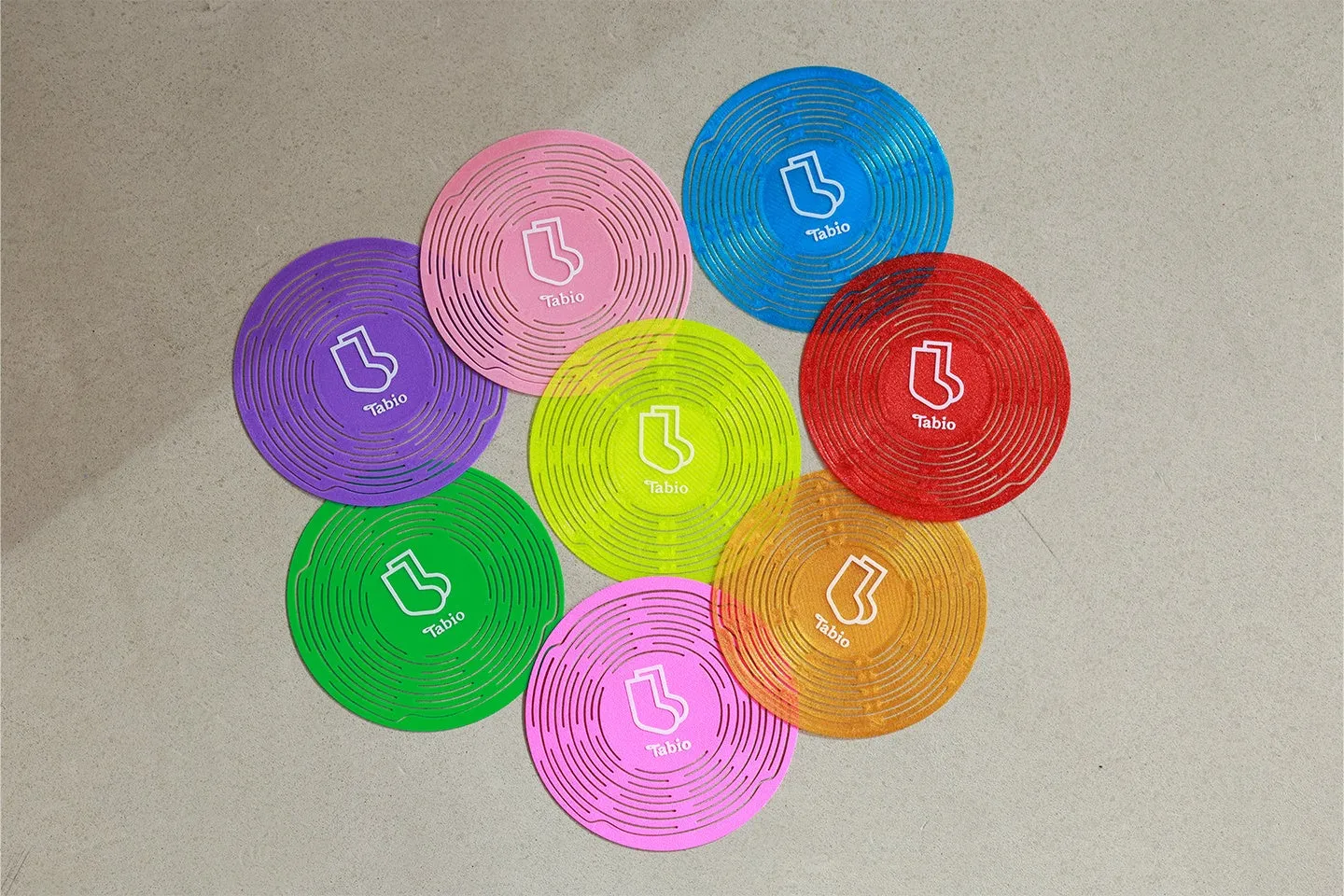 カラフルな円形のコースターが7つ、円形に配置されている画像です。各コースターには、靴下のマークと「Tabio」の文字が白で描かれています。コースターは、紫、ピンク、青、赤、黄緑、オレンジ、緑の7色です。