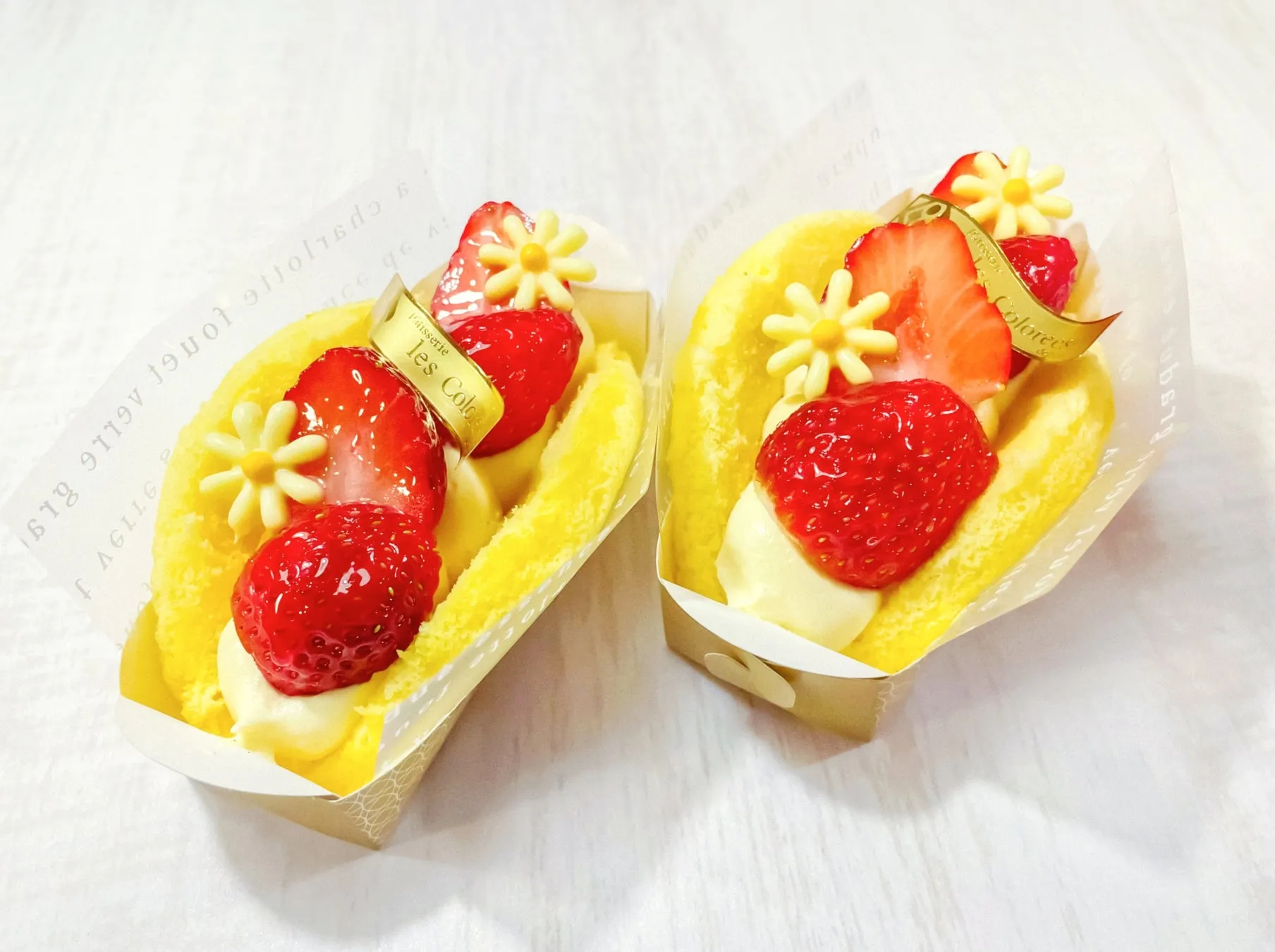いちごがトッピングされた、黄色いケーキが2つ並んだ写真です。ケーキにはクリームが塗られ、花のような飾りと「les Colore」と書かれたリボンが付いています。ケーキは紙の容器に入っており、白い木製のテーブルの上に置かれています。