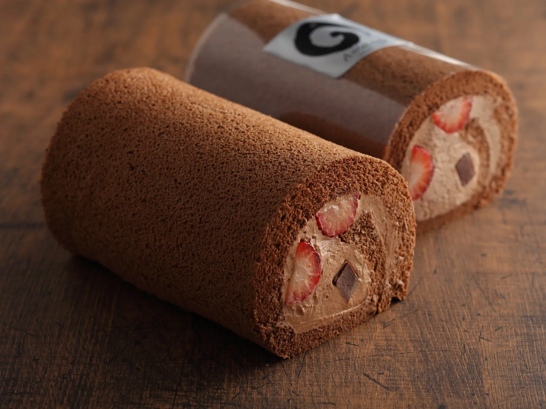 チョコレートロールケーキが2つ並んだ写真です。手前のロールケーキはカットされており、断面にはいちごがトッピングされています。奥のロールケーキは透明なフィルムに包まれています。