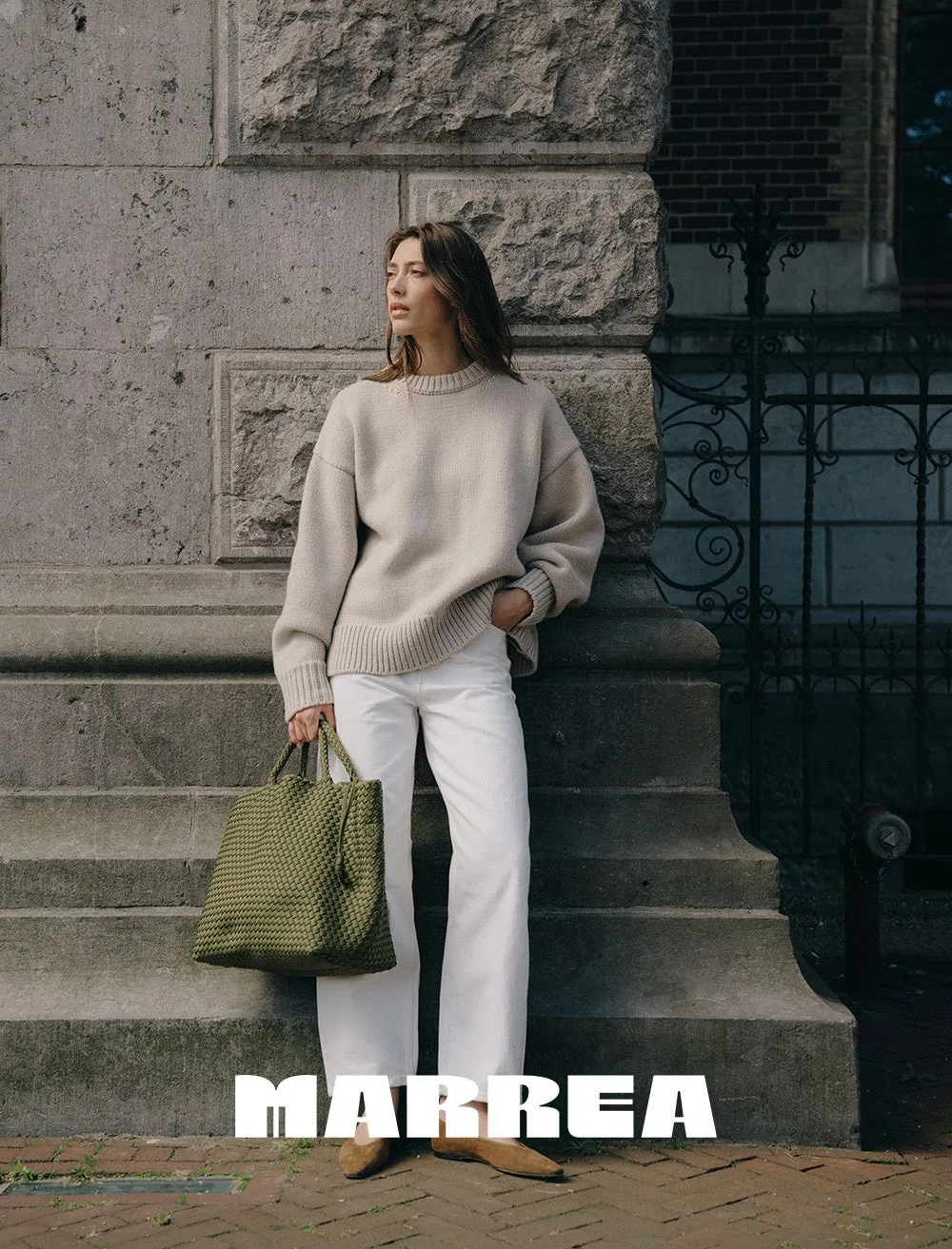 女性が白いパンツとベージュのセーターを着て、緑色のバッグを持っています。彼女は建物の階段に寄りかかっており、ロゴが画像の下部に表示されています。