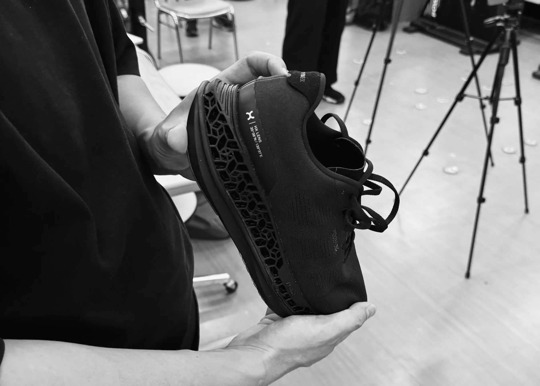 黒いスニーカーを手に持った人のモノクロ写真。スニーカーには、ブランド名と緯度経度が表示されている。