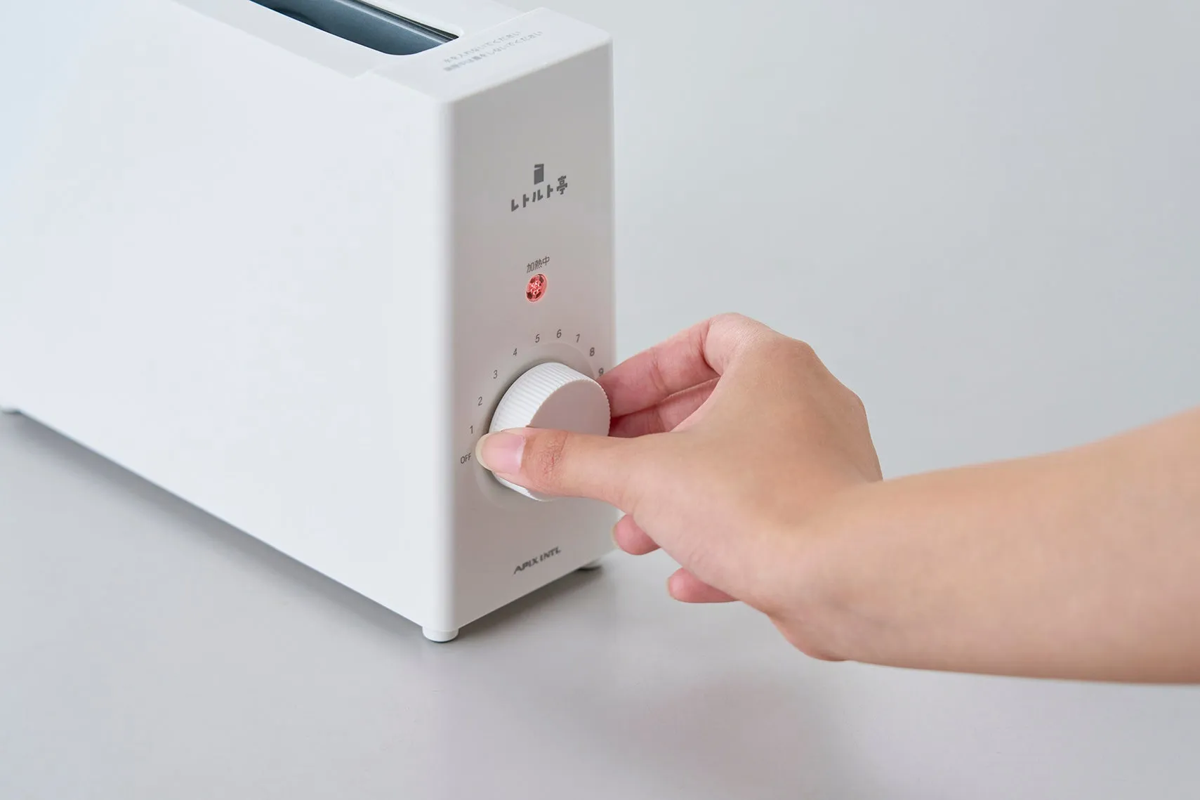 白いトースターのつまみを回している手のクローズアップ。トースターには様々な数字と「OFF」の文字が表示されています。背景はシンプルで、トースターに焦点を当てています。