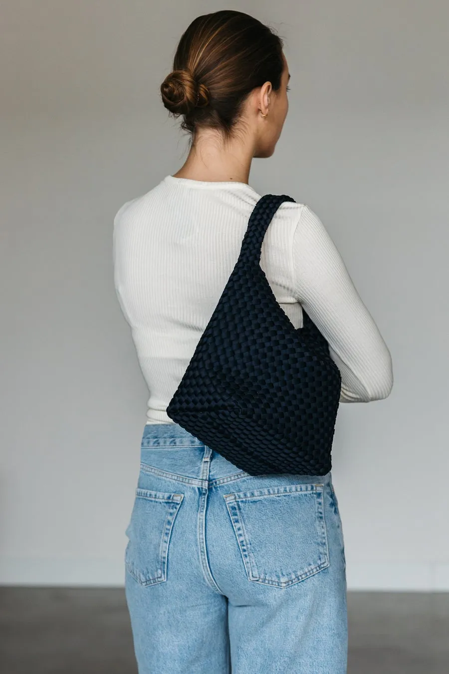 女性が肩に黒色のバッグをかけている写真。女性は白い長袖のシャツとジーンズを着用しており、髪はアップスタイルにまとめられています。