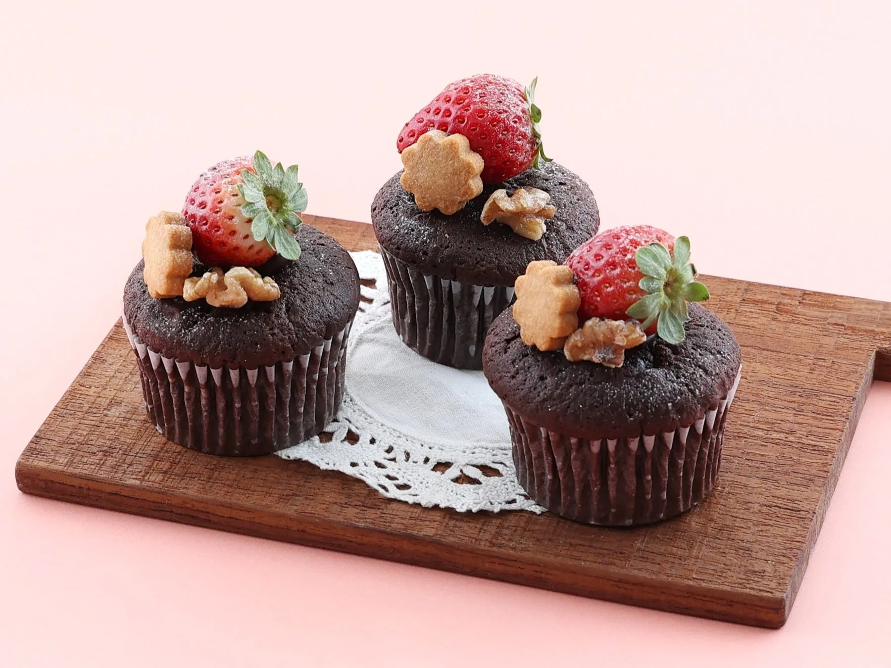 チョコレートカップケーキが3つ、木製のまな板の上に置かれています。それぞれのカップケーキには、イチゴ、クッキー、クルミがトッピングされています。背景はピンク色です。