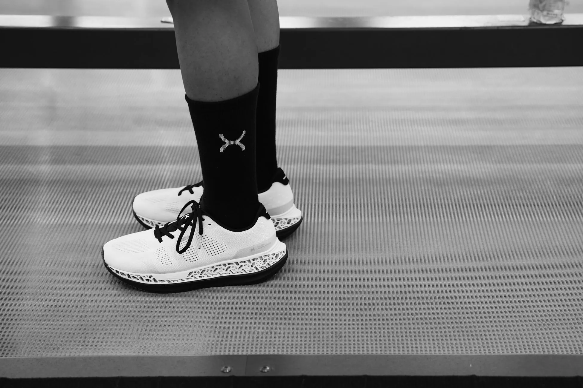 黒い靴下と白いスニーカーを履いた人の足元のモノクロ写真。靴下には白いXのマークがあり、スニーカーは黒い紐で結ばれている。背景はグレーの床。