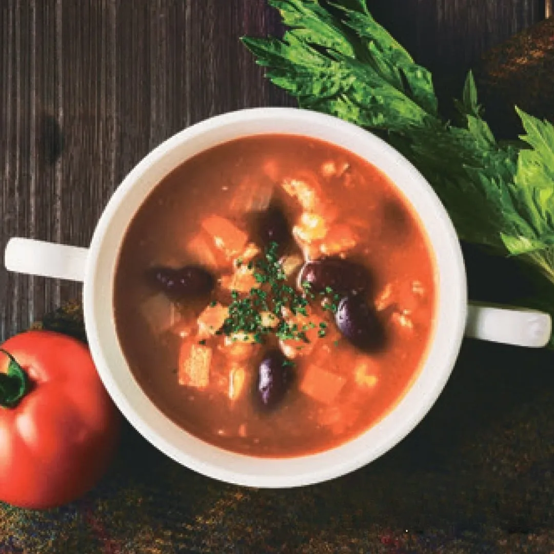 白い器に入ったスープの写真です。スープには、豆や野菜、ハーブが入っています。トマトが添えられています。