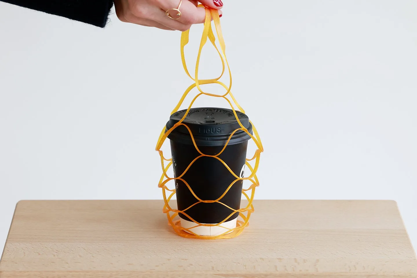 黒いカップに入ったコーヒーを、オレンジ色の網状の持ち手で持っている様子が写っています。手はリングをしていて、背景は白です。